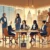 A group of women in business attire engage in discussion around a table in a modern office with large windows, bookshelves, and a cityscape view.